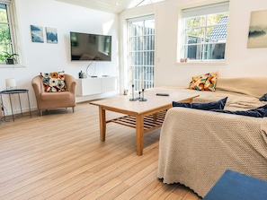 A modern living room with a wooden floor, a sofa, a chair, a TV, and a window with a view of a house.