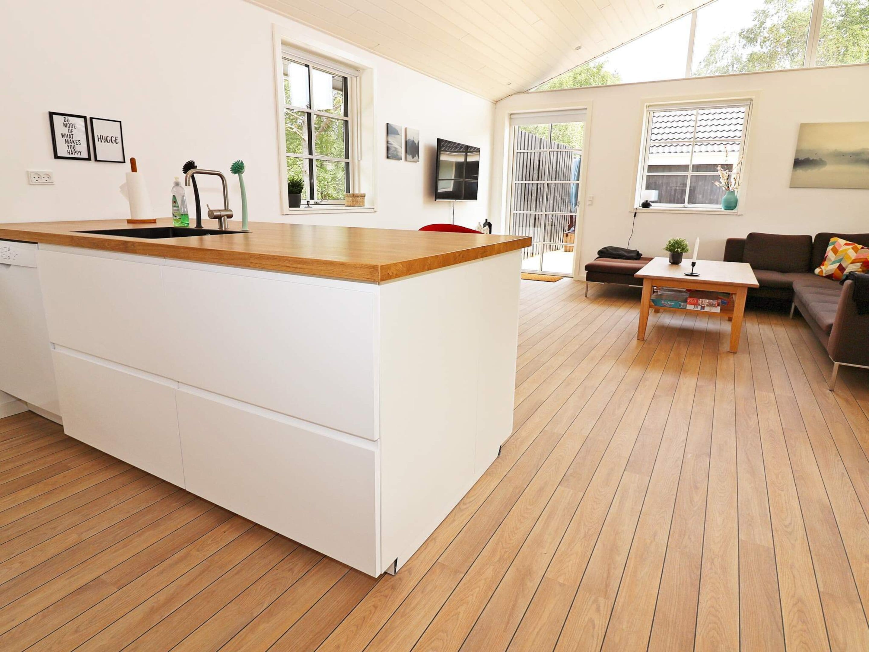 A modern kitchen with a wooden countertop and white cabinetry, a dining area with a wooden table and a sofa, and a large window providing natural light.