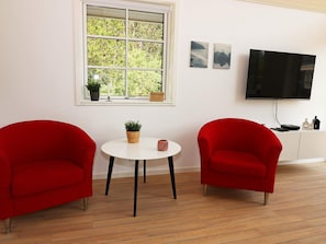 A room with a white wall, a window with a view of trees, two red chairs, a small table with a plant, and a television on the wall. - 8 Person Holiday Home in Hadsund (Hadsund)