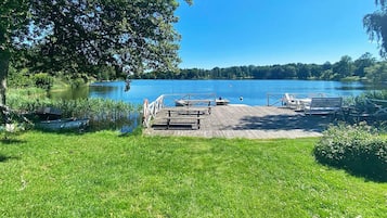 A lakeside deck with picnic tables and chairs, surrounded by lush greenery and a clear blue sky.