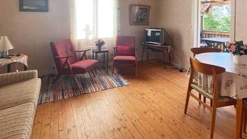 A room with wooden flooring, a sofa, two armchairs, a small table, a television, and a window with a view of greenery.