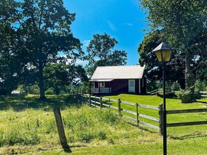 A small, single-story building with a porch, surrounded by a wooden fence and lush greenery. - 4 Person Holiday Home in Vetlanda (Vetlanda)
