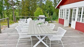 A wooden deck with a dining table and white wicker chairs, a barbecue grill, and a red building with white windows in the background.