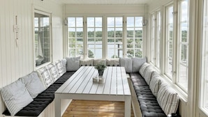 Uma sala de sol com interior branco, janelas amplas, uma mesa de madeira e um sofá com padrão preto e branco.