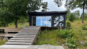Uma cabana moderna com um deck de madeira que leva a uma porta de vidro, rodeada por vegetação natural.