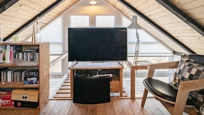 A cozy attic room with a large TV, wooden bookshelves, a comfortable chair, and a view through a large window. - 2 Person Holiday Home in Brovst (Brovst)