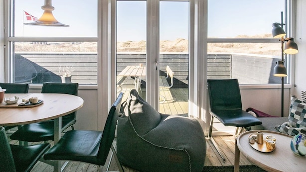 A cozy room with a wooden ceiling, large windows, and a view of the beach. There are chairs, a table, and a lamp. - 2 Person Holiday Home in Brovst (Brovst)