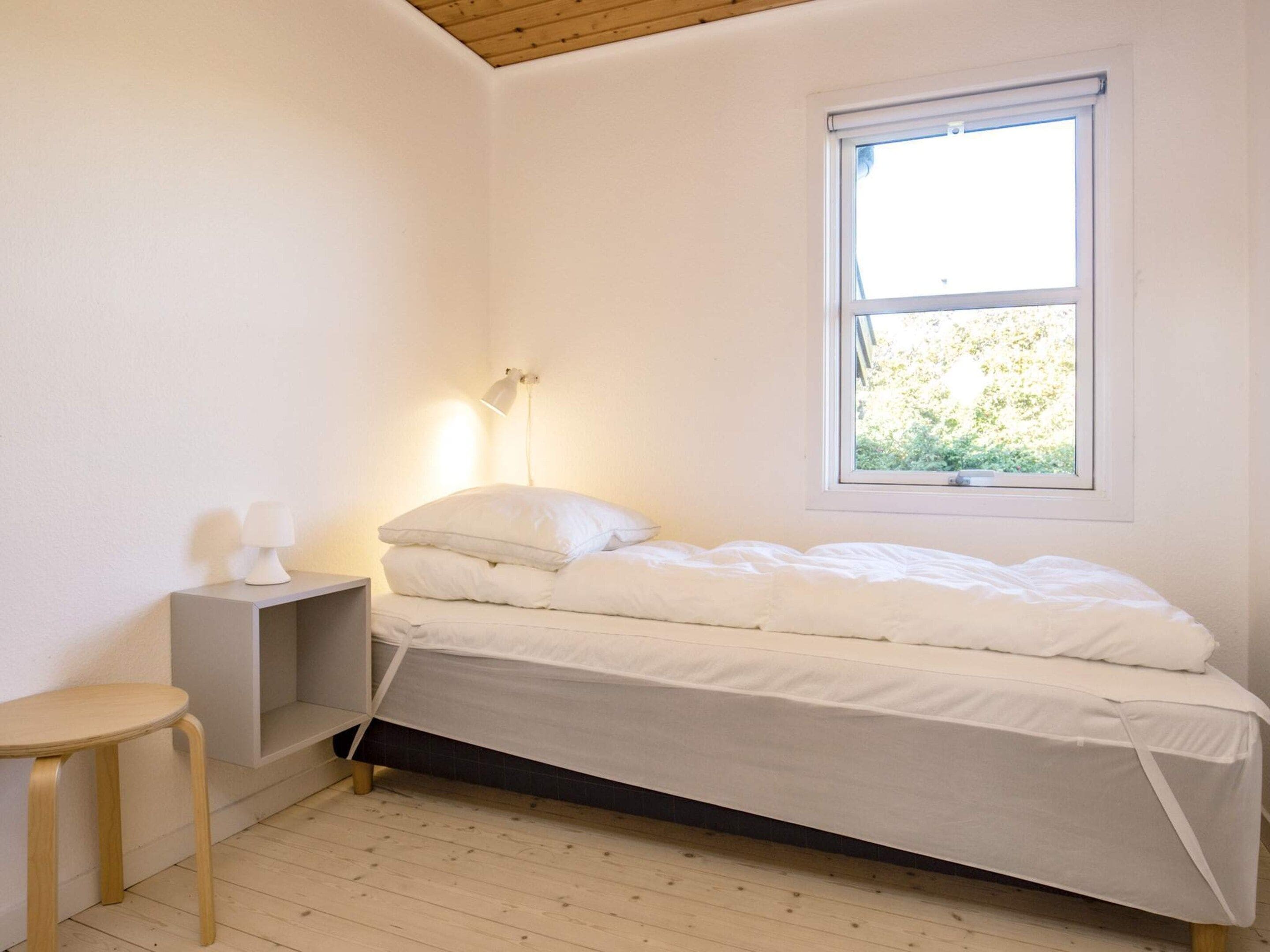 Cama pequena e bem arrumada, com roupa de cama branca, em quarto com paredes brancas e piso de madeira. Tem uma mesinha de cabeceira, uma luminária e um banquinho pequeno. Uma janela com vista para o verde é visível.