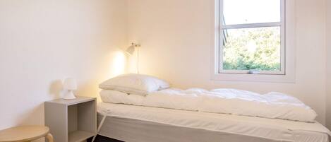 A small, neatly made bed with white bedding in a room with white walls and a wooden floor. There is a bedside table with a lamp and a small stool. A window with a view of greenery is visible.
