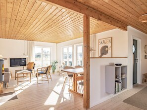 A spacious living area with a wooden ceiling, a dining table, and chairs. There is a television mounted on the wall, a bookshelf, and a window providing natural light. - 4 Person Holiday Home in Skagen (Skagen)