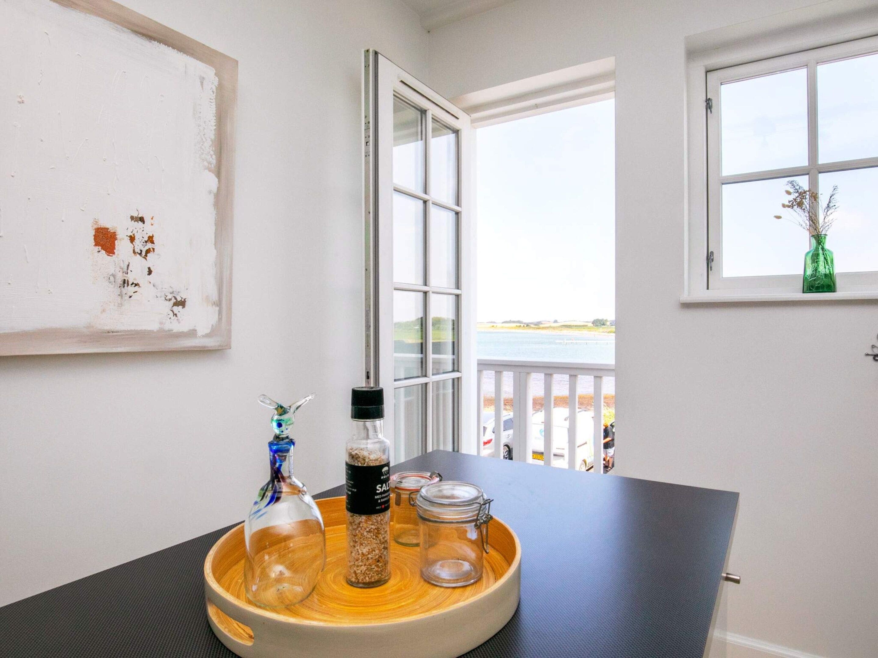 Une pièce contient une table sur laquelle se trouvent une bouteille en verre, un bocal et un récipient. Une fenêtre donne sur un plan d’eau et on aperçoit un balcon.