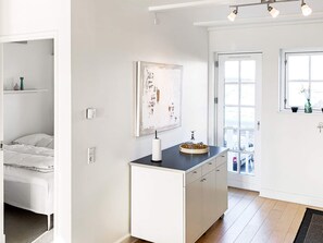 Un intérieur moderne avec un lit, une commode blanche à tiroirs, un tableau au mur et une fenêtre donnant sur un paysage.