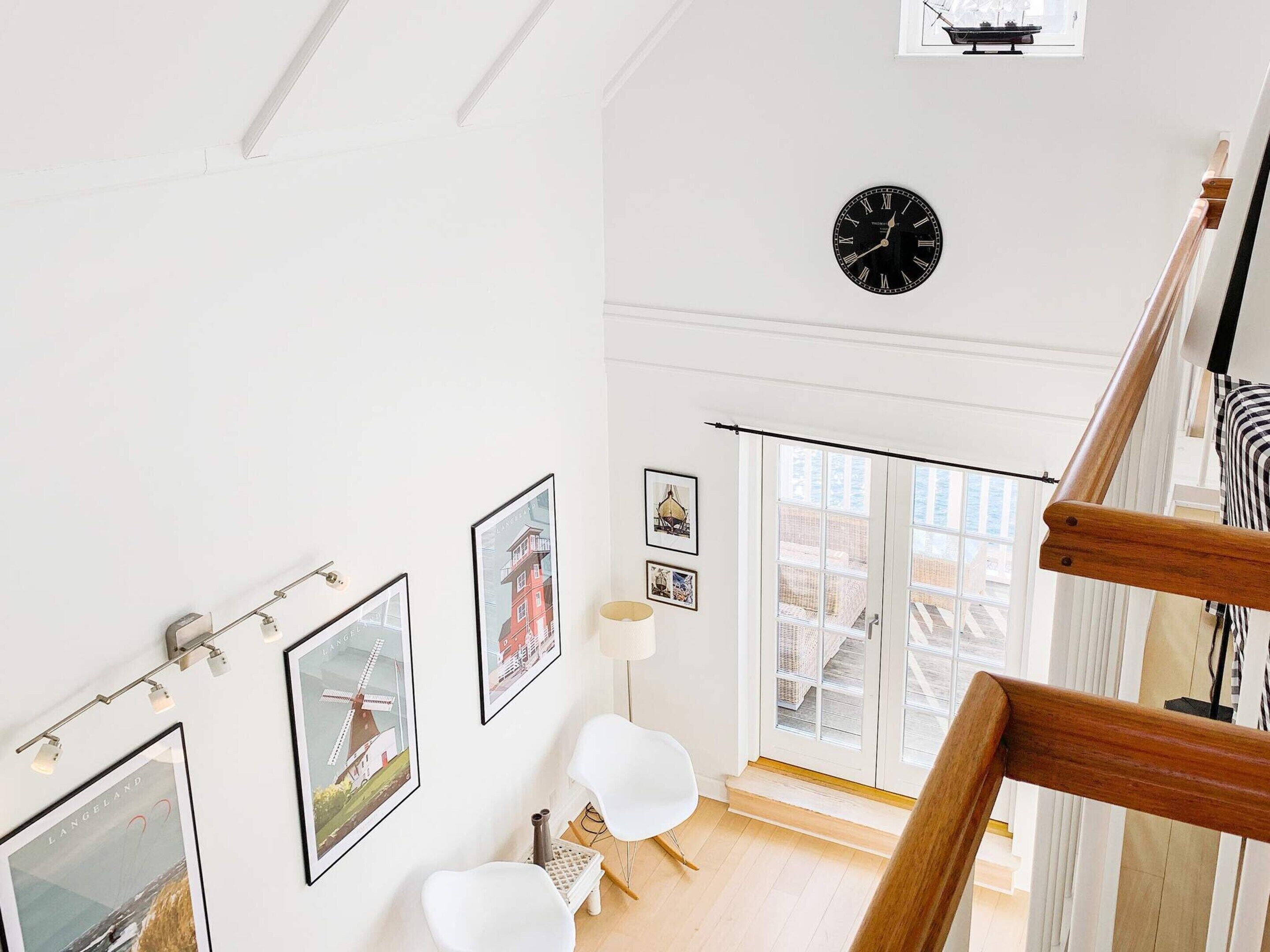 A spacious room with a wooden staircase, a black clock on the wall, and framed pictures on the walls.