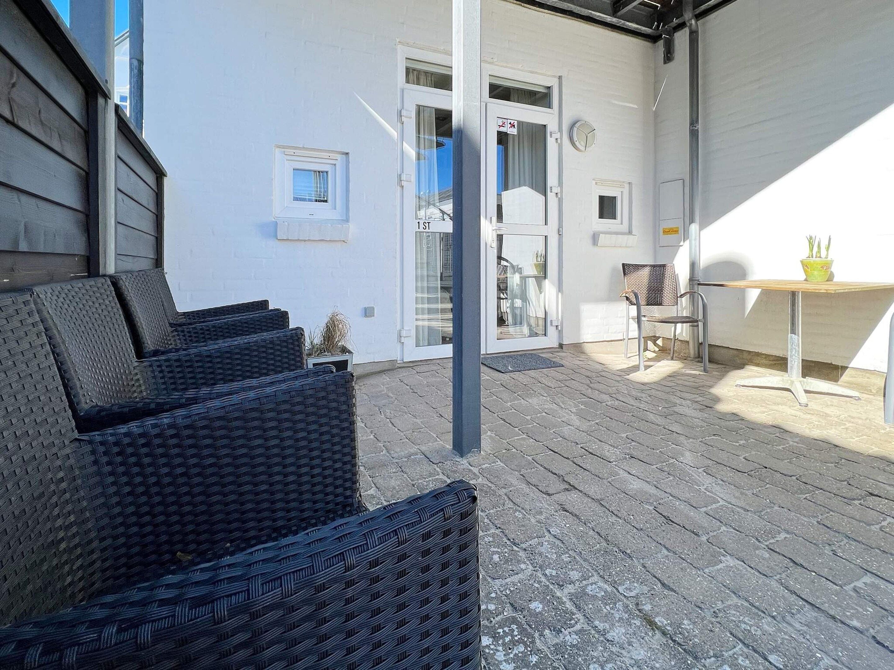 A covered outdoor seating area with wicker furniture, a small table with a bowl, and a door leading to a building.