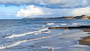A coastal scene with waves, a sandy beach, and a rocky cliff with buildings. - 4 Star Holiday Home in Lokken (Lokken)
