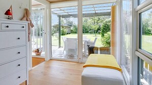 Una habitación con techo de madera, un aparador blanco con cajones, un banco con una manta amarilla y un amplio ventanal que ofrece vistas a un jardín y mobiliario exterior.