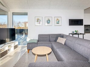 A modern living room with a grey sofa, a round coffee table, and a flat-screen TV. There are three framed artworks on the wall and a kitchen area with a black TV and cabinets. - 4 Star Holiday Home in Vinderup (Vinderup)