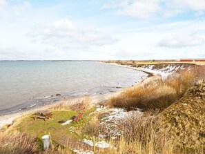 A coastal landscape with a sandy beach, a grassy area with a bench and picnic table, and a red building in the distance. - 4 Star Holiday Home in Vinderup (Vinderup)