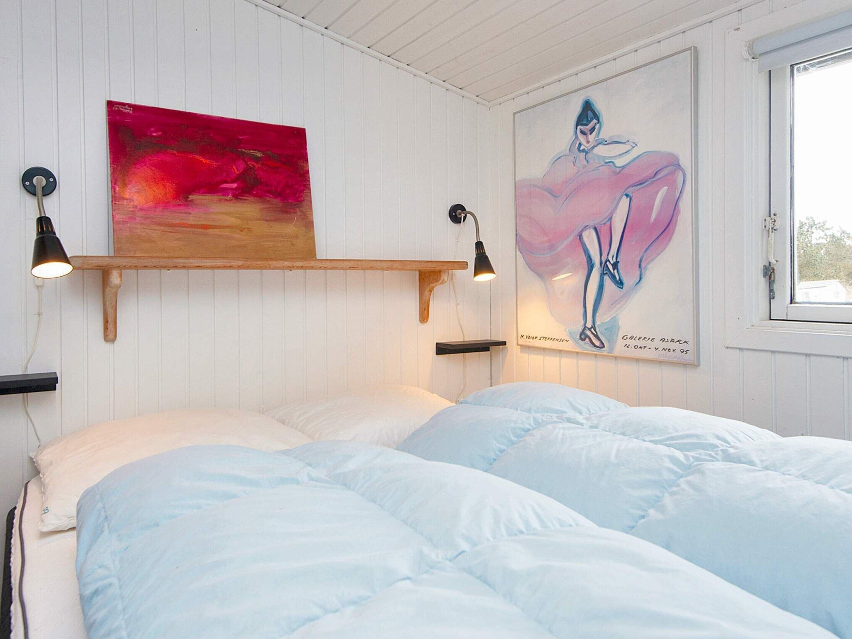 Um quarto com uma cama, duas luminárias na parede, uma prateleira de madeira com um quadro e uma janela.