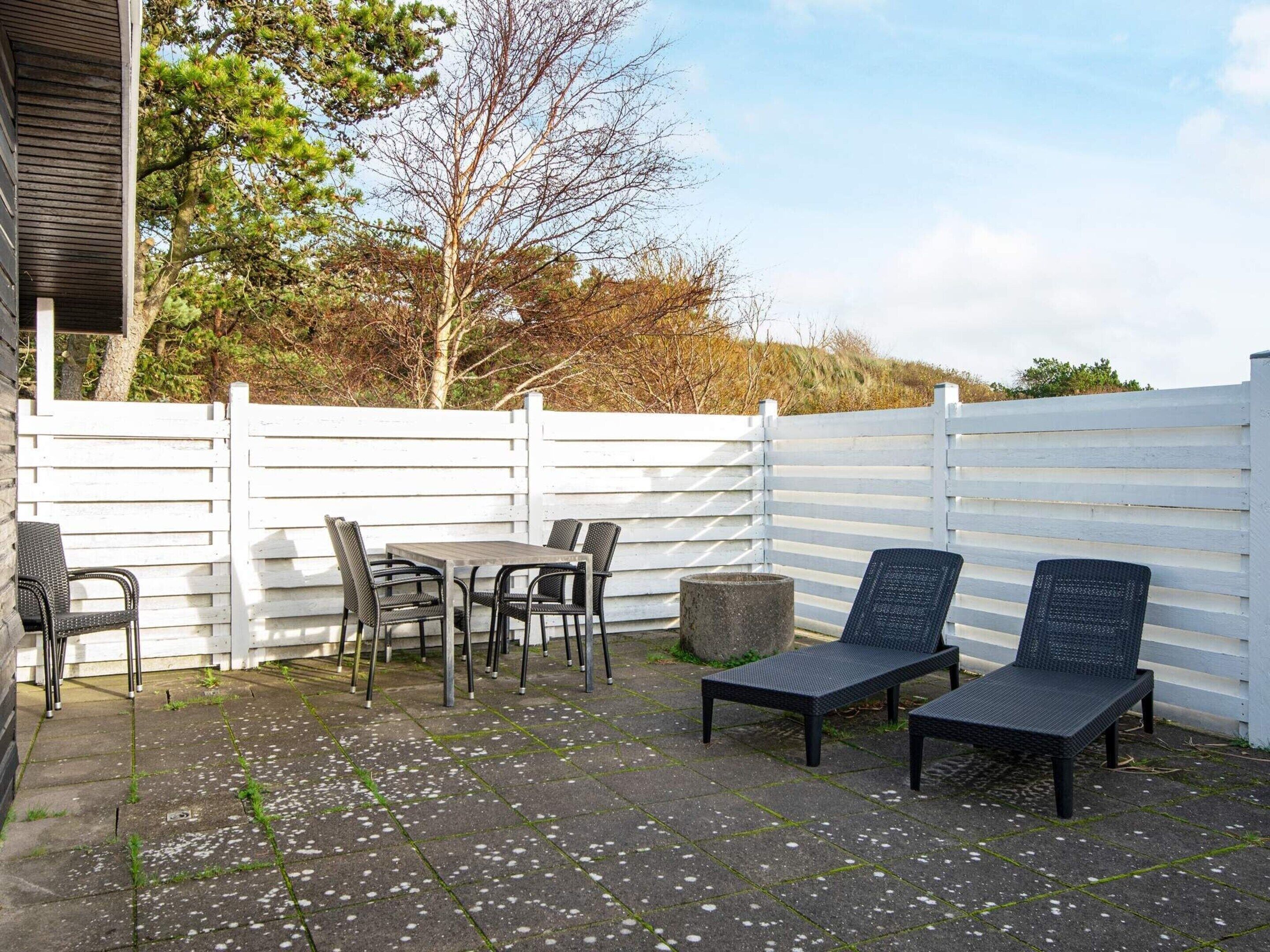 Une terrasse extérieure avec une table et des chaises, un jardinier en pierre et une clôture blanche.