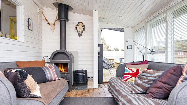 A cozy living room with a wood-burning stove, two grey sofas with patterned cushions, and a large window with blinds. - 6 Person Holiday Home in Fano (Fano)