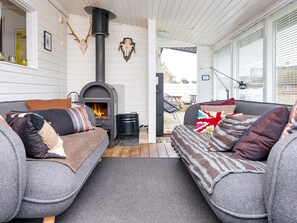 Ein gemütliches Wohnzimmer mit Kaminofen, zwei grauen Sofas mit gemusterten Kissen und einem großen Fenster mit Jalousien.