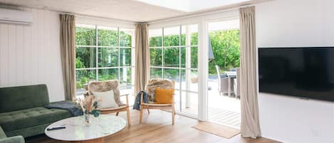 Une pièce avec un canapé vert, une table basse en verre, deux fauteuils, un téléviseur et une grande fenêtre donnant sur un paysage verdoyant.