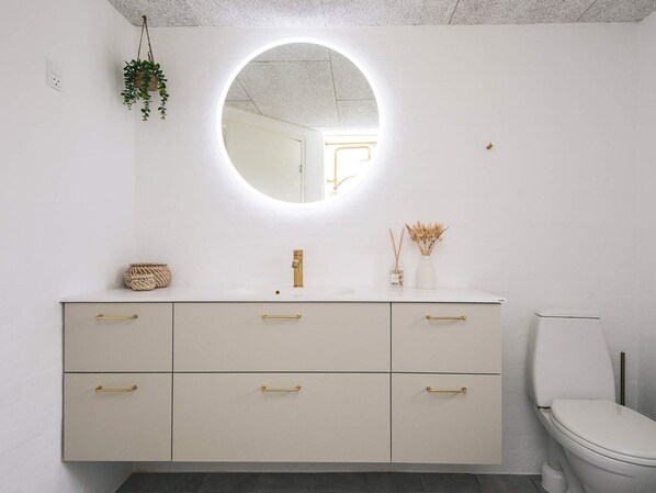 Une salle de bain moderne avec un meuble-lavabo blanc, des poignées dorées, un miroir rond avec éclairage intégré et des toilettes.