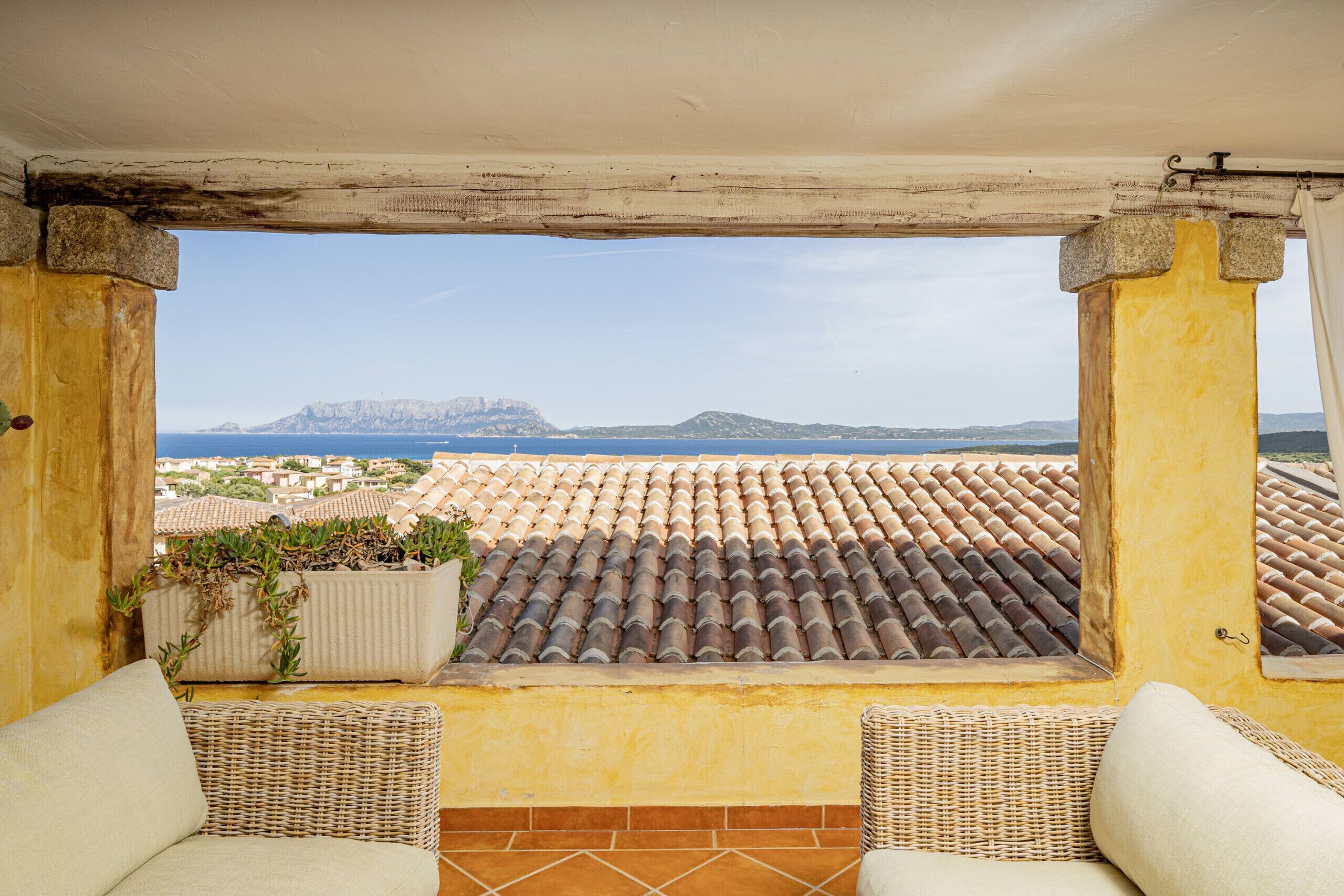 A balcony with a view of the sea and mountains, wicker furniture, and terracotta tiles.