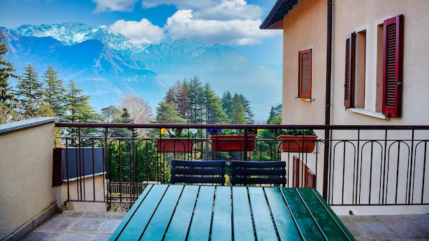 Ein Balkon mit Blick auf schneebedeckte Berge, einem Holztisch und roten Fensterläden an einem Gebäude.