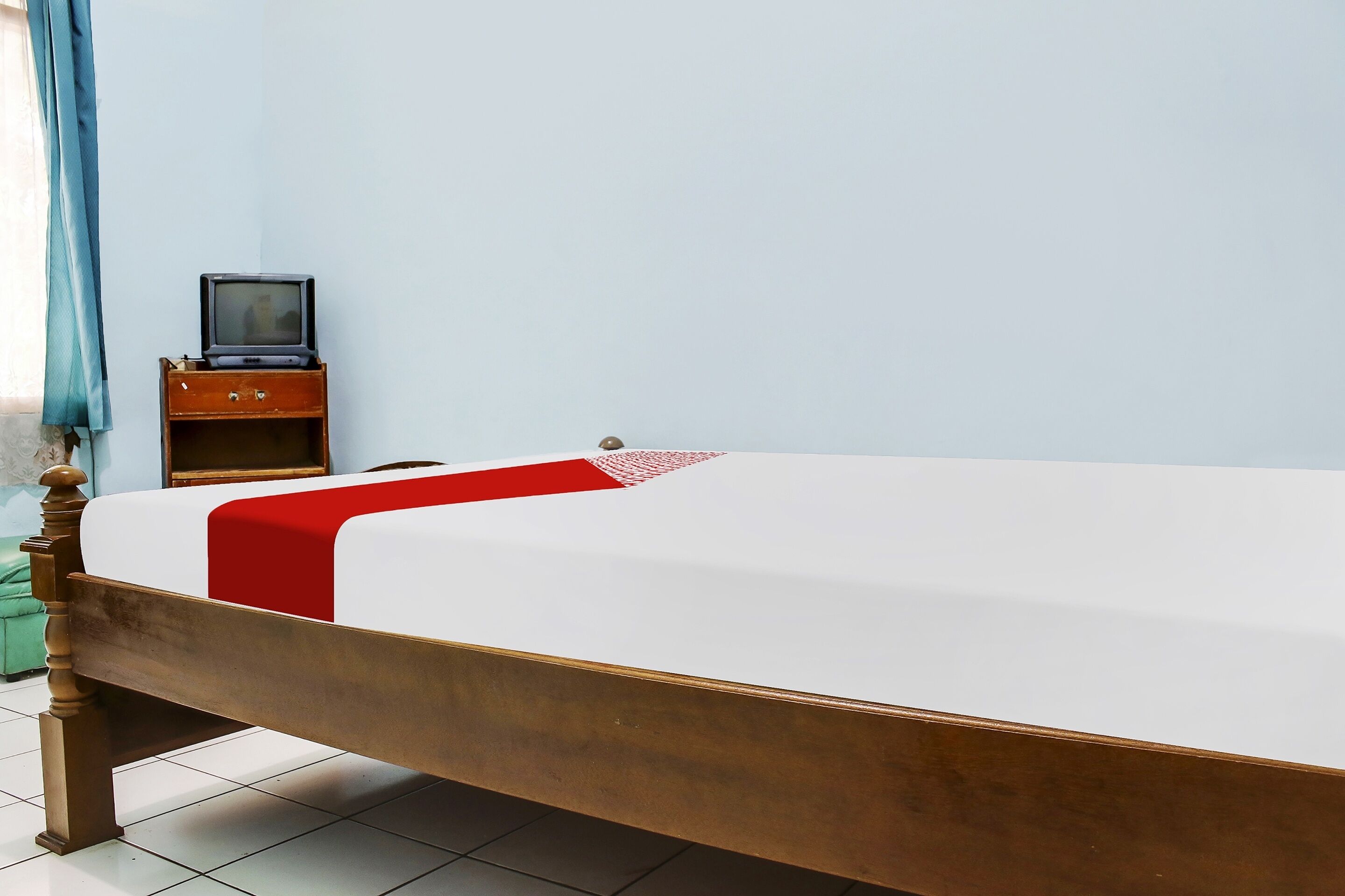 Een bed met houten frame en een wit matras, een rode deken en een klein nachtkastje met een televisie.
