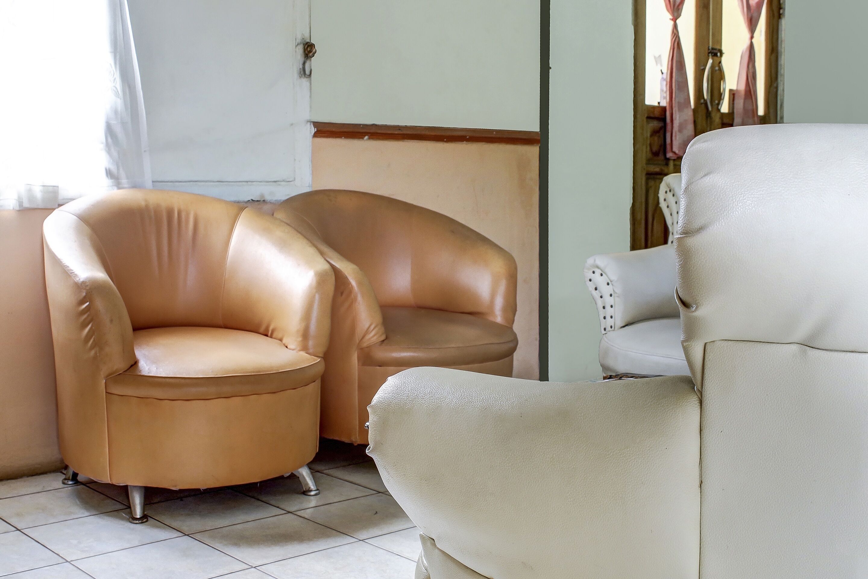 A room with two leather armchairs and a white armchair, a wooden door with a handle, and a tiled floor.