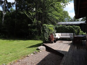 A wooden deck with a white outdoor sofa and a table, surrounded by greenery and trees.