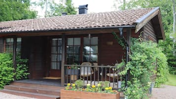 A wooden cabin with a tiled roof, a porch with a railing, and a garden with flowers.