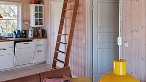 A cozy room with a wooden interior, a sofa with cushions, a yellow lamp, and a kitchen area with cabinets and a ladder. - 5 Person Holiday Home (Vordingborg)