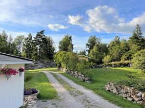 A gravel path leading to a white building with flower baskets, surrounded by a well-maintained lawn and stone walls, under a partly cloudy sky. - 2 Person Holiday Home in Henan (Henån)