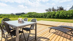 Une terrasse en bois à l’extérieur, avec une table et des chaises, entourée de verdure et sous un ciel dégagé.