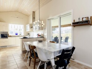 A modern kitchen with a dining area, featuring a large island, white chairs, and a view through a sliding glass door. - 6 Person Holiday Home in Nysted (Nysted)
