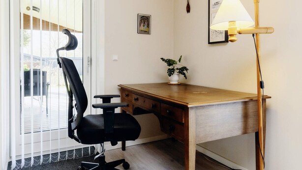 A small office with a wooden desk, a black office chair, a potted plant, a wall-mounted lamp, and a door with blinds. - 6 Person Holiday Home in Nysted (Nysted)