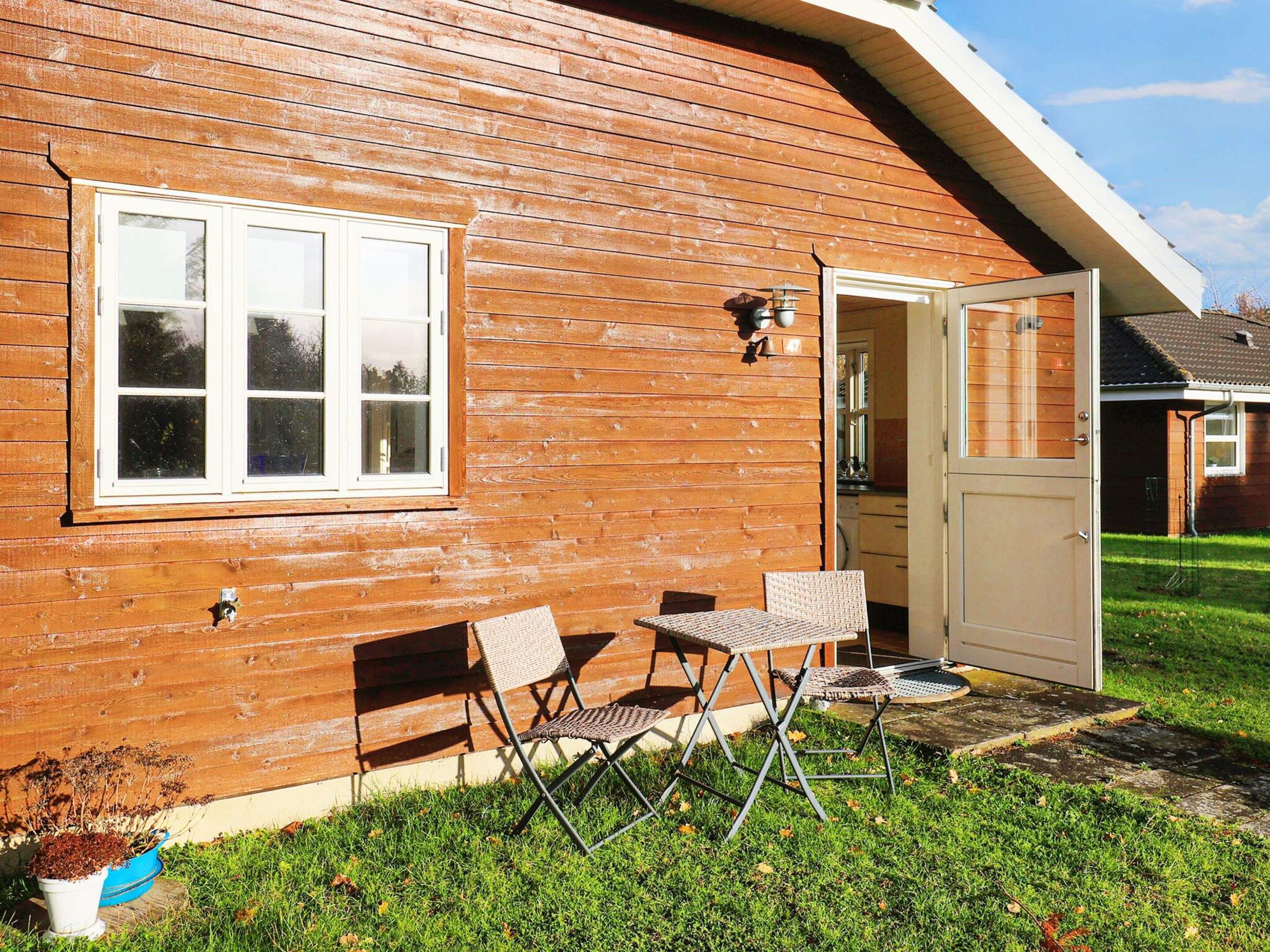 Une cabane en bois avec une fenêtre à cadre blanc, une porte et une petite terrasse avec deux chaises et une table.