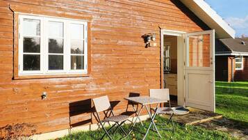 A wooden cabin with a white-framed window, a door, and a small patio with two chairs and a table.