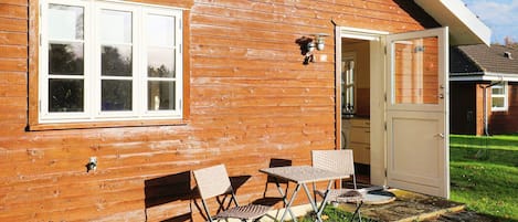 A wooden cabin with a white-framed window, a door, and a small patio with two chairs and a table.