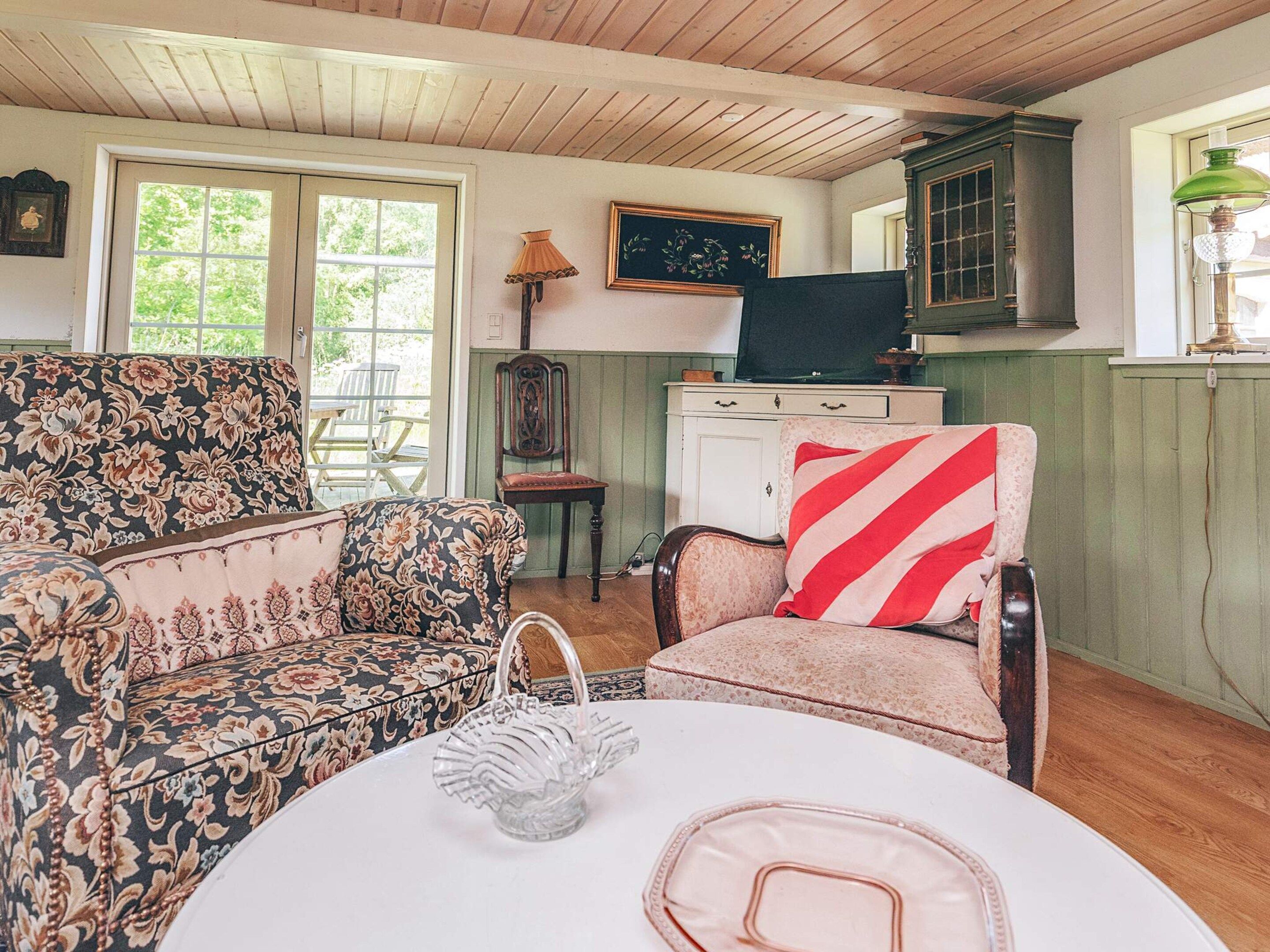 A cozy room with a floral-patterned sofa, a chair with a striped pillow, a small table with a vase, and a cabinet with a television.