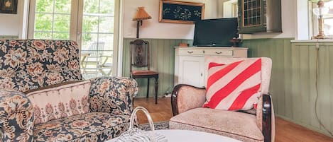 A cozy room with a floral-patterned sofa, a chair with a striped pillow, a small table with a vase, and a cabinet with a television.