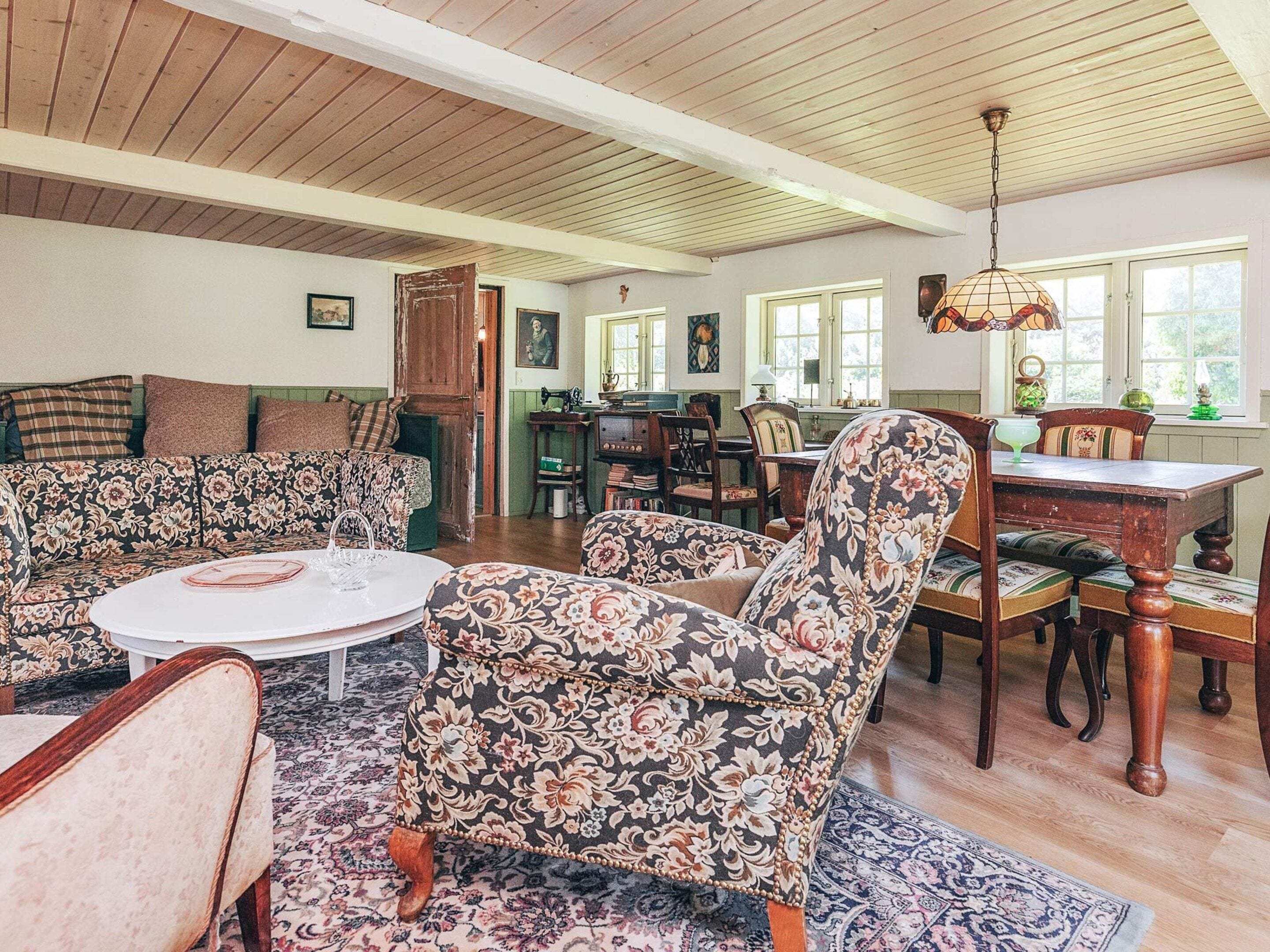 A spacious living room with a high ceiling, floral-patterned armchairs, a round coffee table, and a dining area with a wooden table and chairs.
