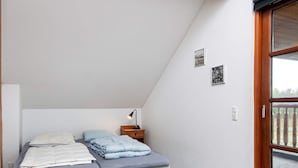 Quarto com teto inclinado, cama com colchão cinza e roupa de cama azul, luminária de cabeceira e janela com vista para uma varanda.