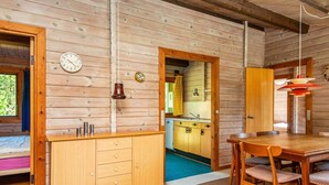A wooden cabin interior with a dining table, chairs, a kitchen area, and a clock on the wall.
