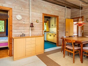 A wooden cabin interior with a dining table, chairs, a kitchen area, and a clock on the wall.
