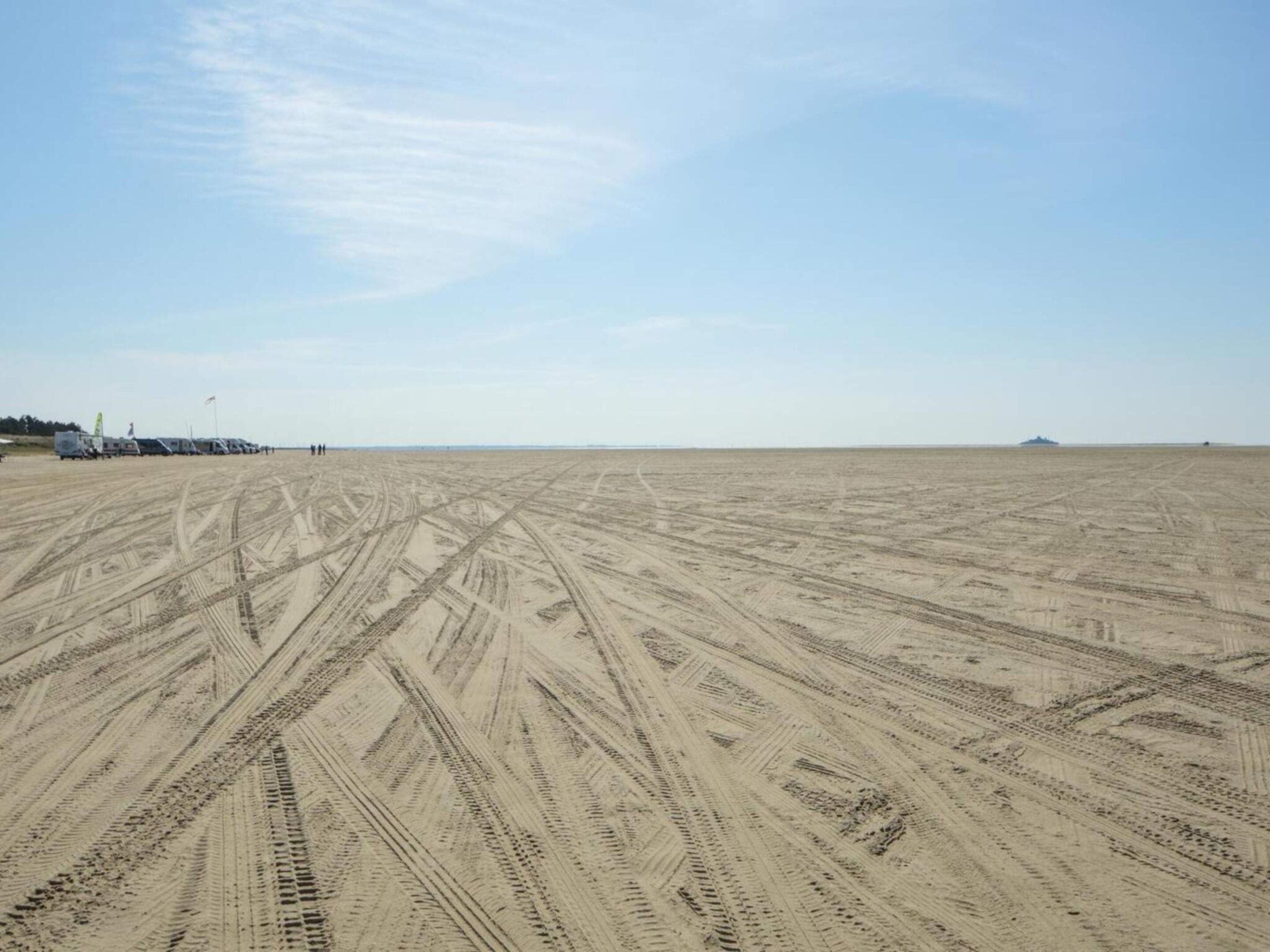 Une vaste étendue sablonneuse, à ciel ouvert, parsemée de traces de pneus, avec un ciel dégagé et des bâtiments visibles au loin.