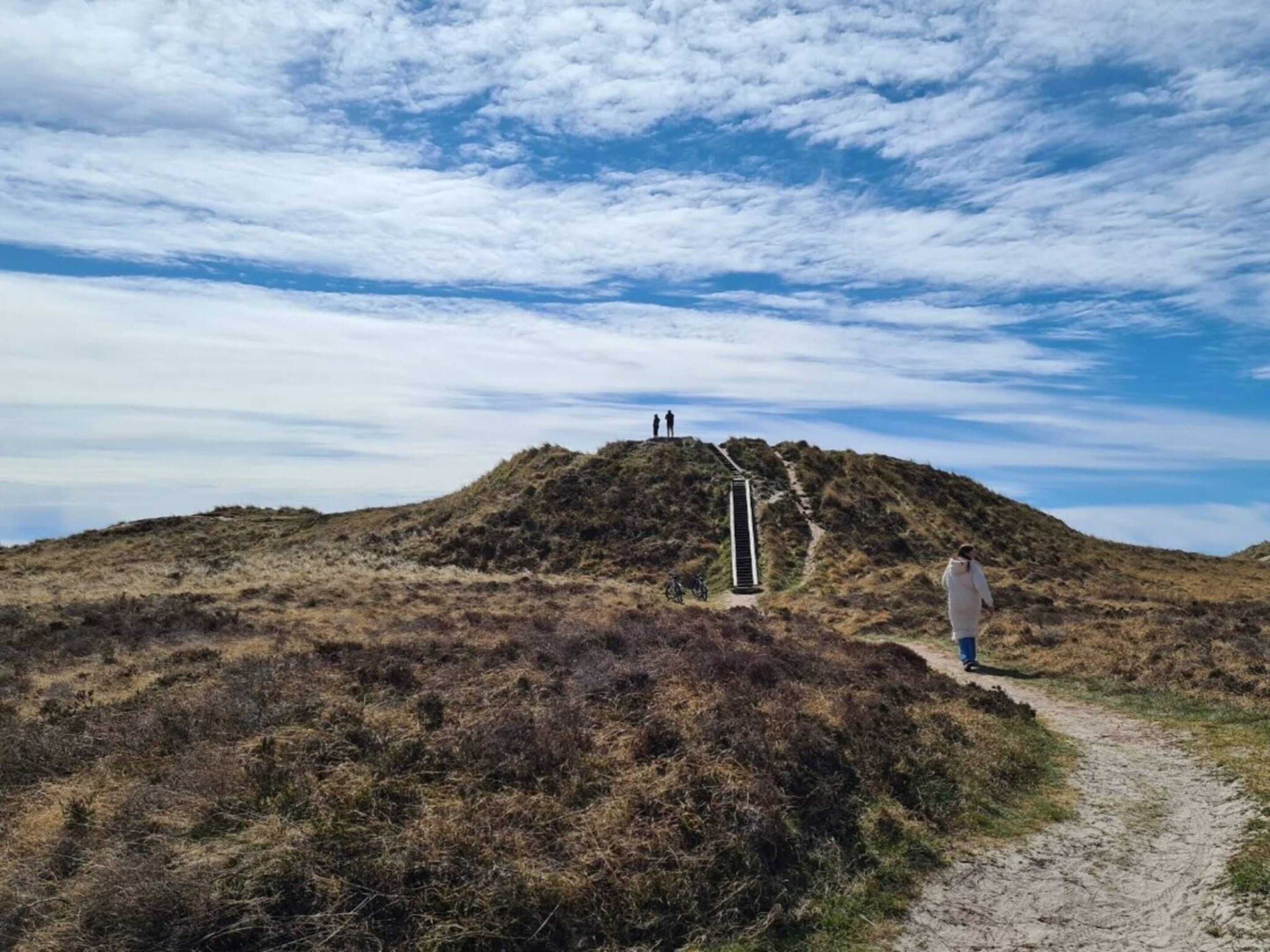 Een persoon loopt over een onverhard pad naar een heuvel met een metalen trap die leidt naar een uitkijkpunt.
